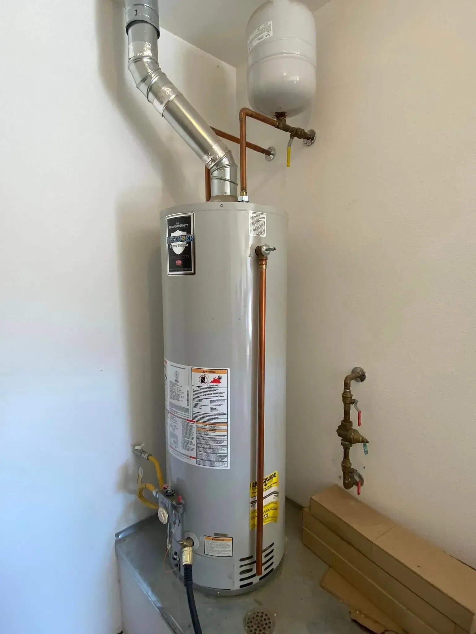 A tall, silver water heater with copper pipes in a white-walled utility room.