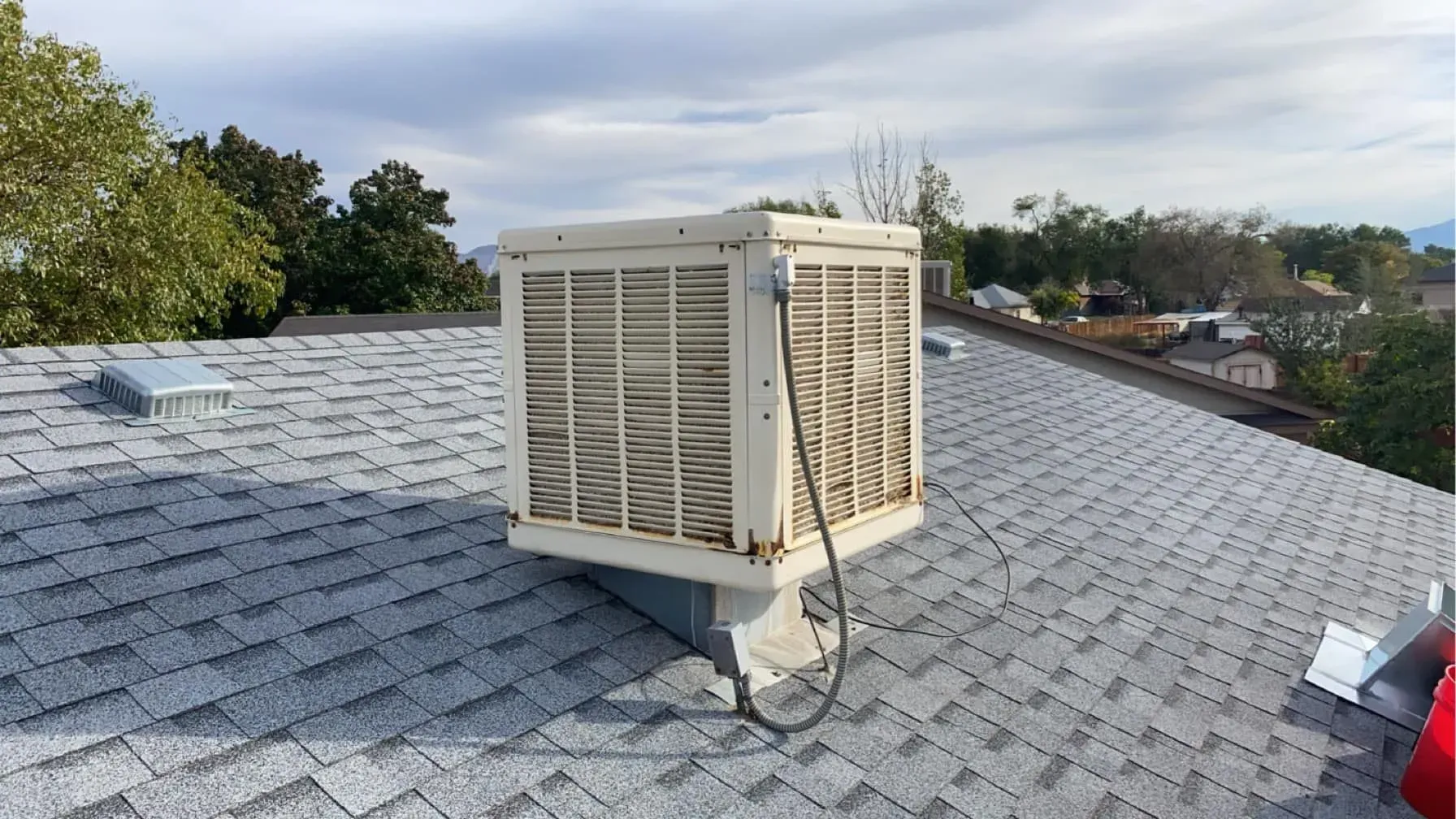 Rooftop with an evaporative cooler; gray shingles, cloudy sky, and trees in the background.