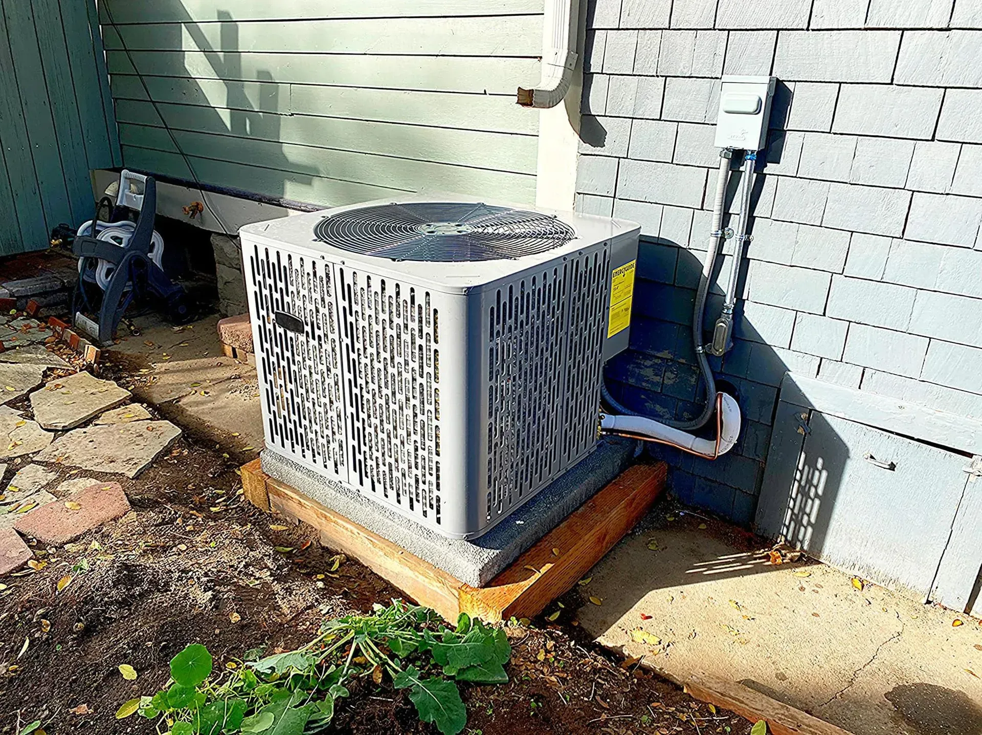 Air conditioning unit outside, gray and square, on a wooden base, near a brick wall and green plant.