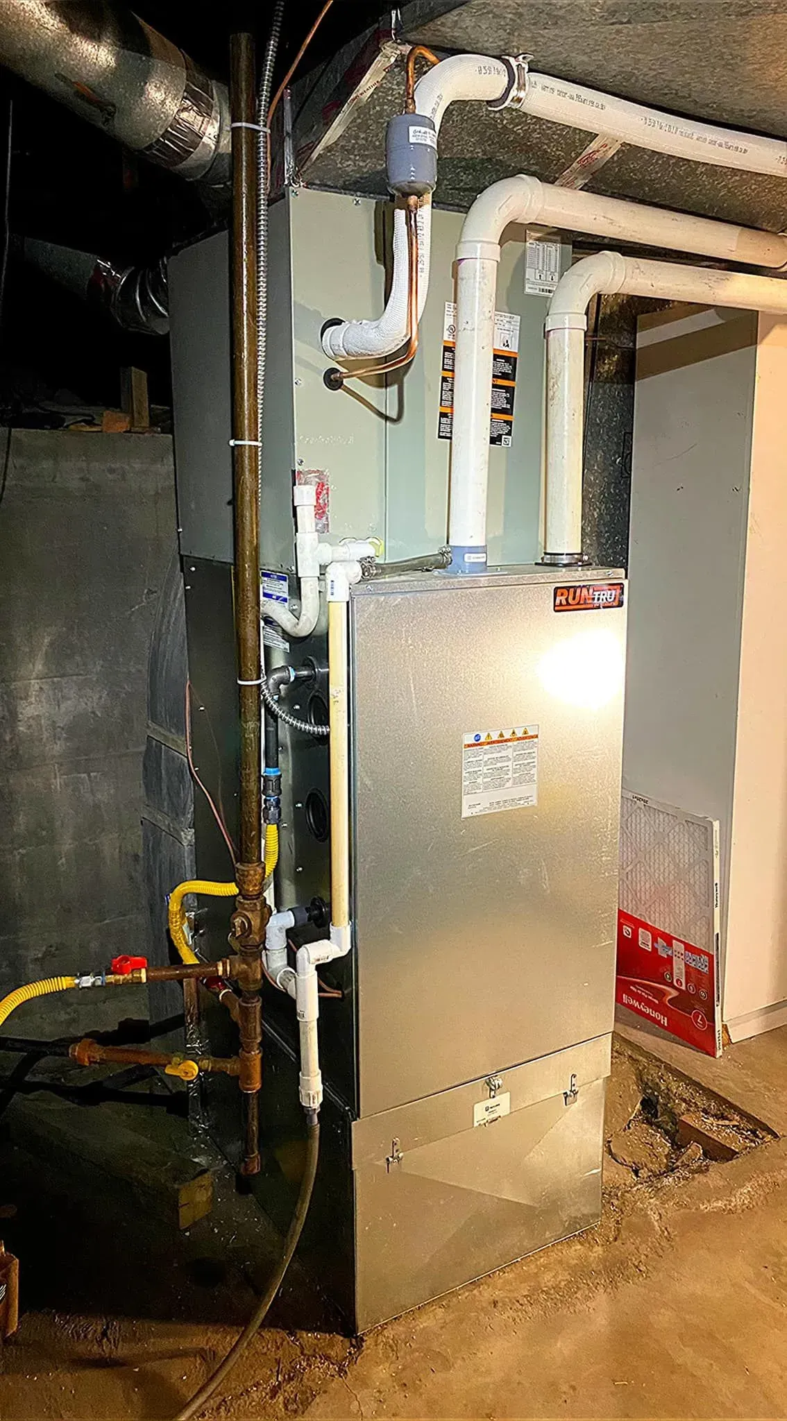 Furnace in a basement with pipes and ductwork. Metallic and white components, on a concrete floor.