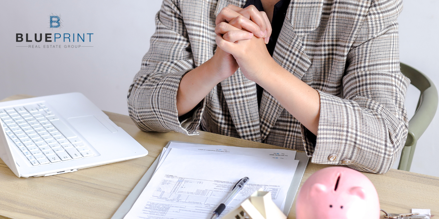 Person with hands clasped over paperwork, small house model, pen, piggy bank, and laptop on desk.