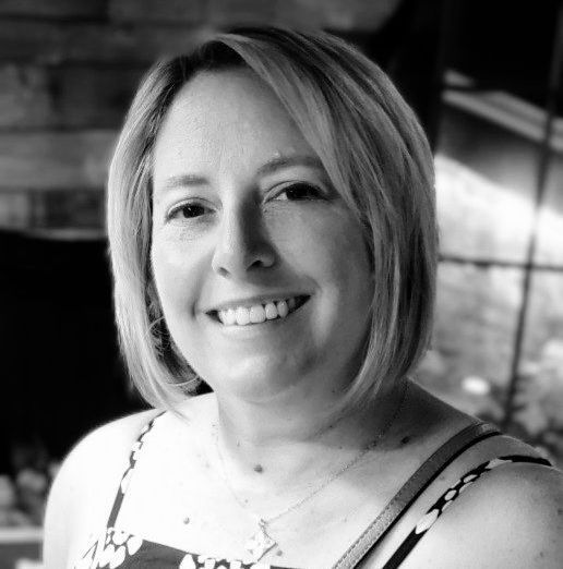 Woman with blonde hair smiles, wearing a patterned top, in an outdoor setting.