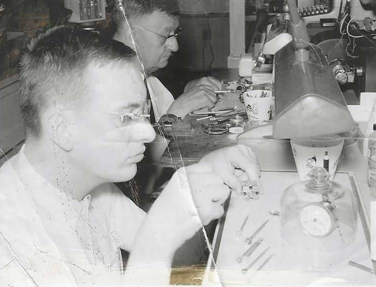 Two people working on small objects at a workbench, likely watch repair.