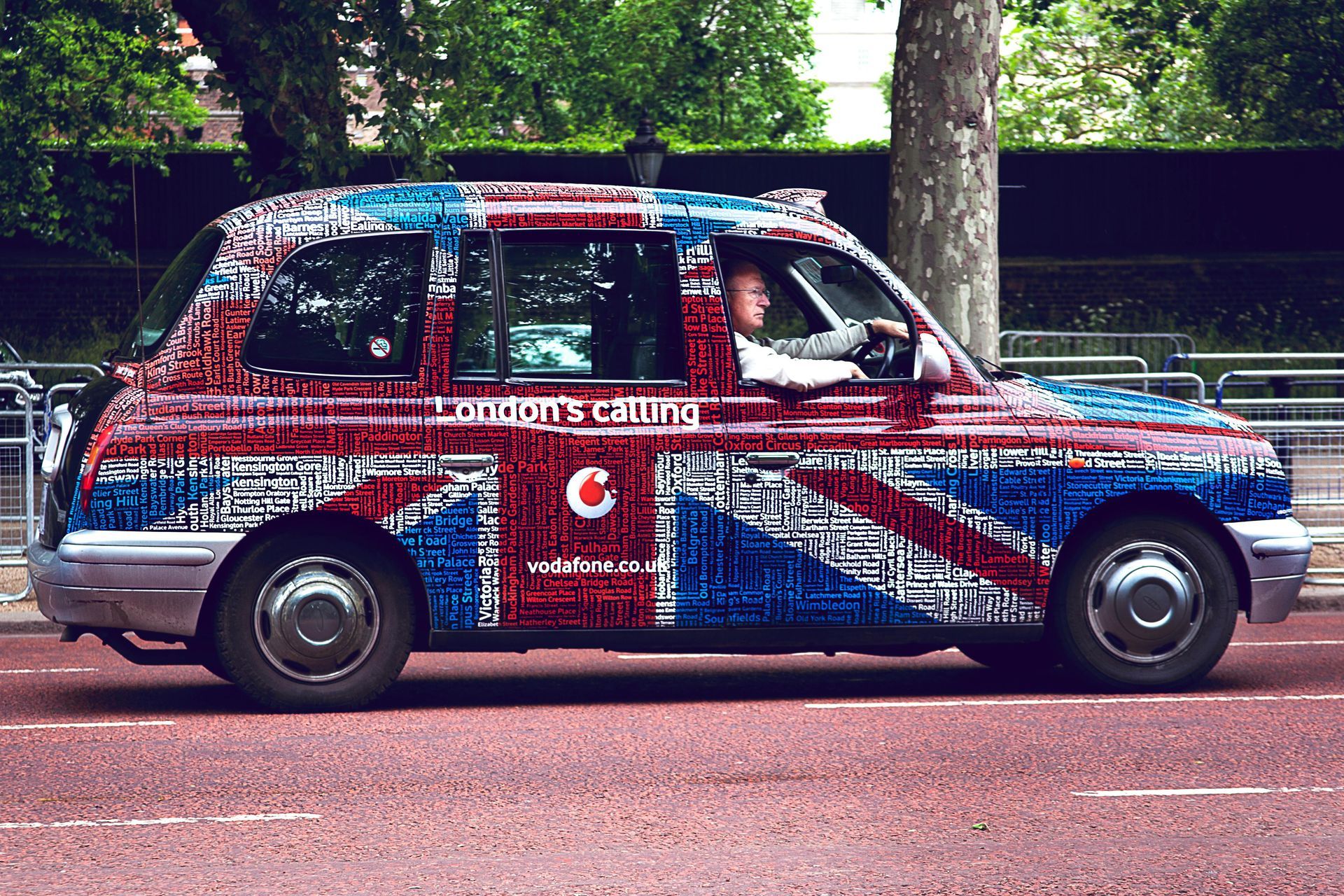 A London taxi cab with a Union Jack design drives on a red road with a driver inside.