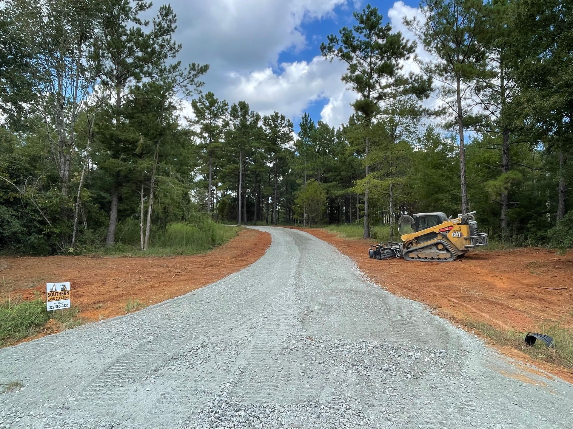 Gravel driveway curves through a wooded area; a bulldozer sits to the side. Blue sky with clouds.