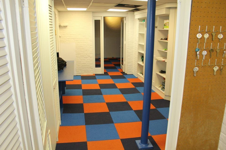 A room with checkered orange, blue, and black floor tiles, shelves, and a blue support pole. Keys hang on the right.