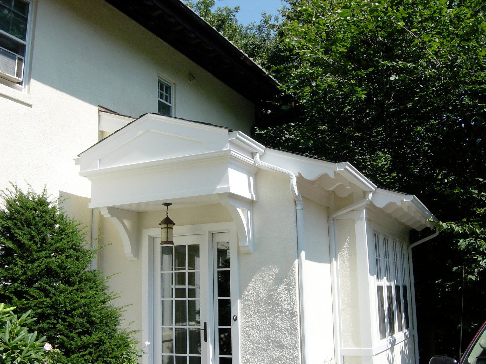 White house with porch and door under a decorative overhang; green trees and bushes surround it.