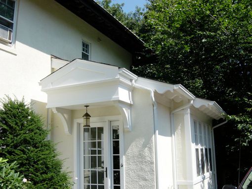 White house with porch and door under a decorative overhang; green trees and bushes surround it.