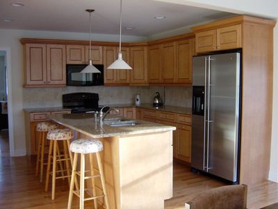 Kitchen with light wood cabinets, granite countertops, stainless steel refrigerator, and a kitchen island with stools.