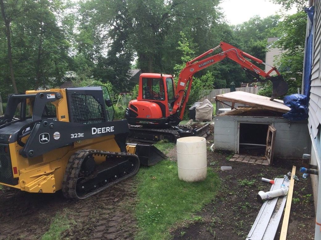 Construction site: excavator and skid steer working near a small building.