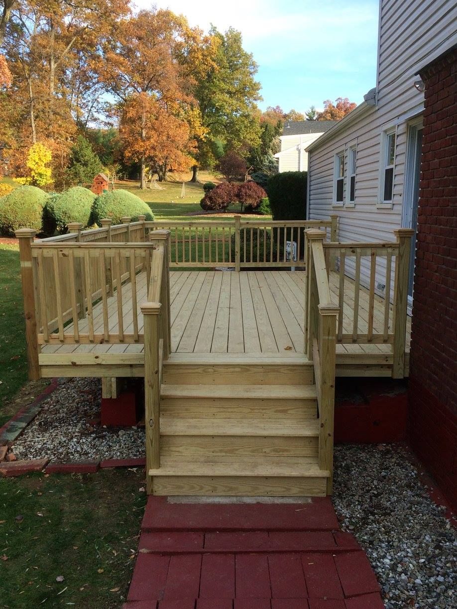 Wooden deck with stairs, railing, and red brick path in front of a house with fall foliage.