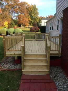 Wooden deck with stairs, railing, and red brick path in front of a house with fall foliage.