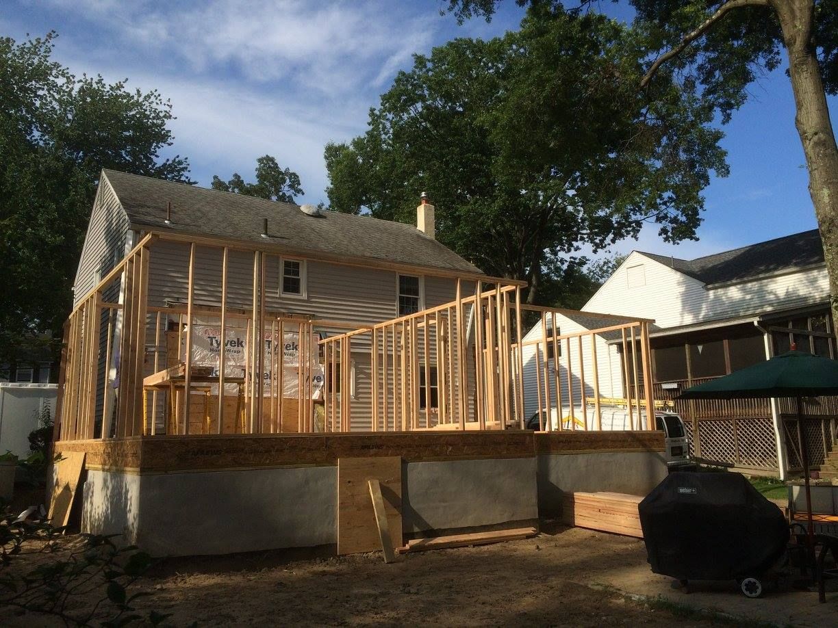 Wooden frame of an addition to a house; under construction with a partial deck, against a background of trees.