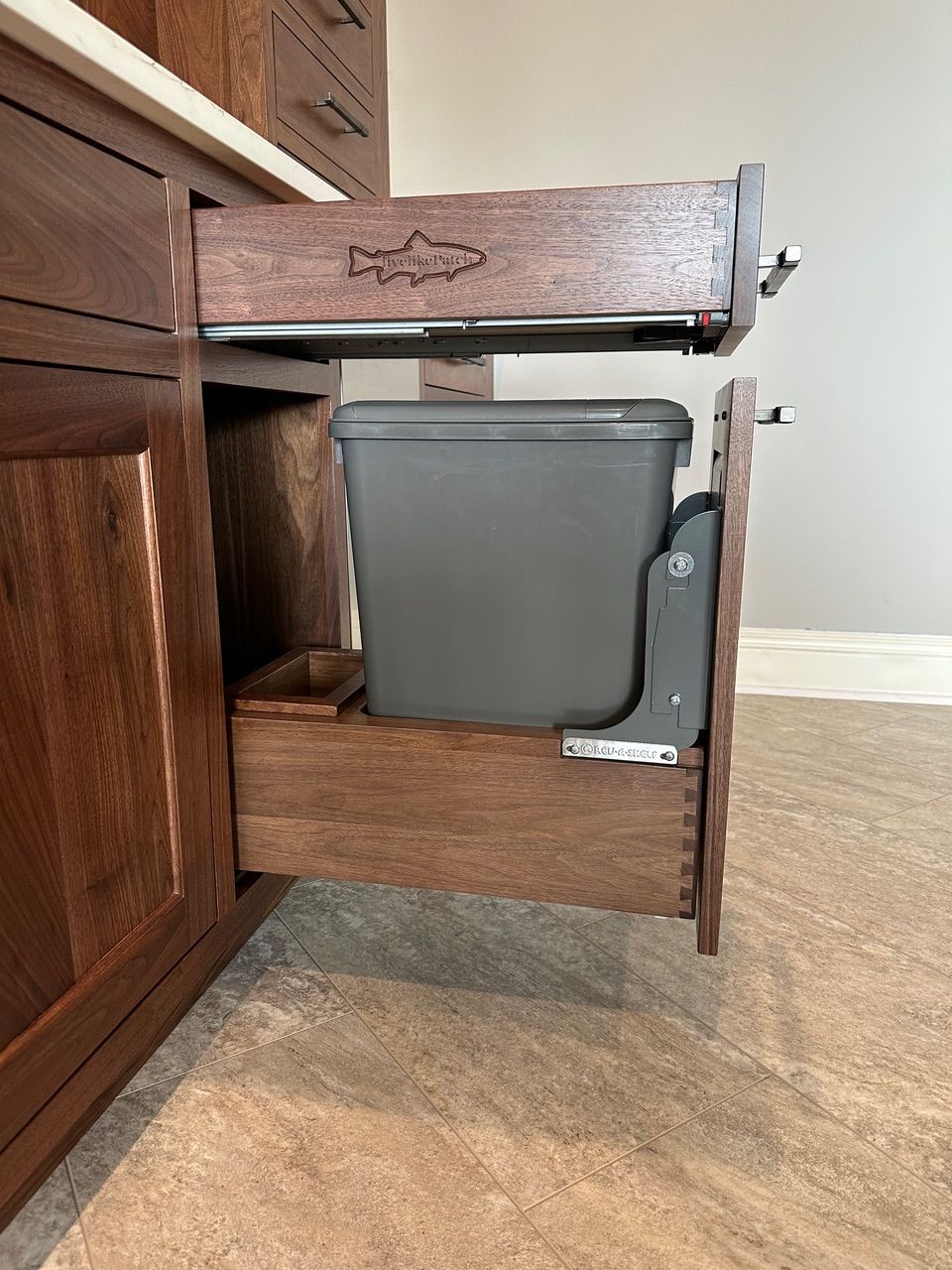 Open wooden cabinet with a pull-out trash can. Dark brown wood, gray trash bin, on a beige floor.