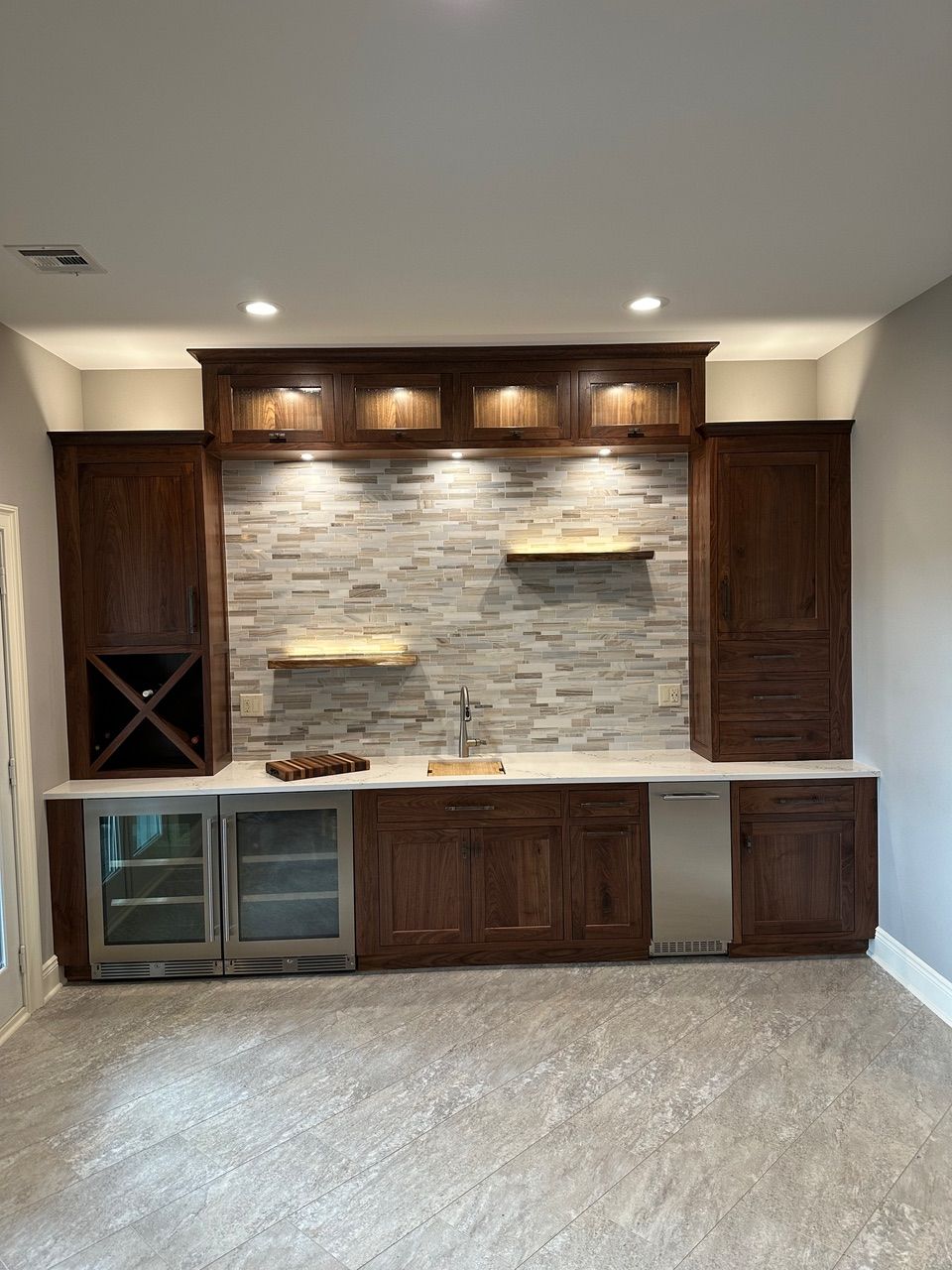 Dark wood home bar with stone backsplash, cabinets, wine cooler, and sink.