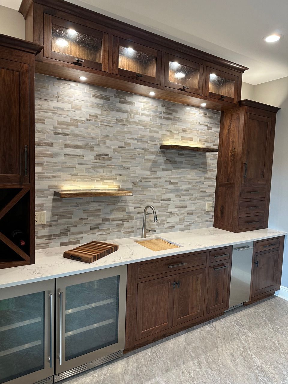 A home bar with wooden cabinets, stone backsplash, and built-in wine and beverage coolers.