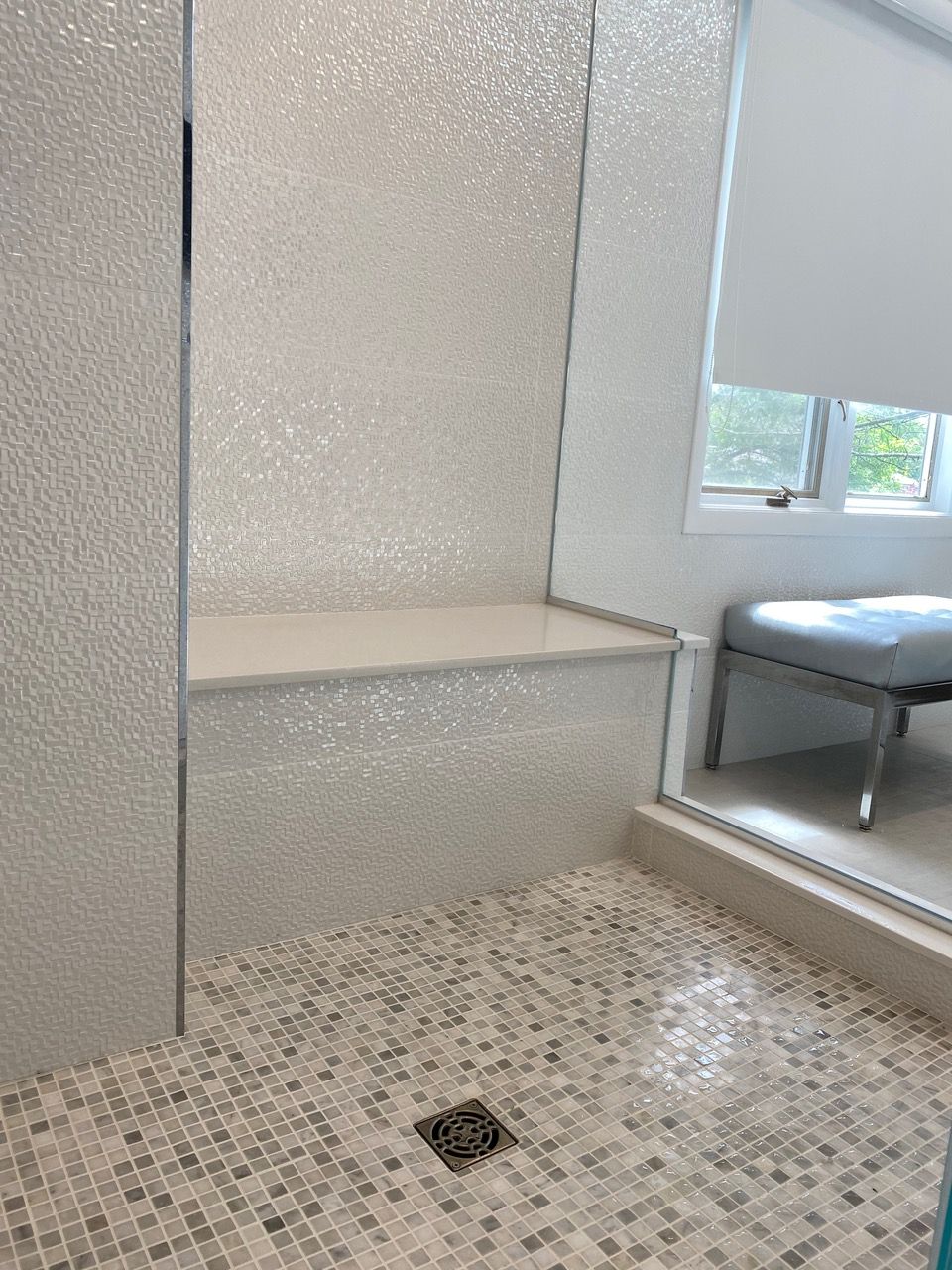 Shower with textured white tile walls, built-in seat, and mosaic floor. A window and bench are visible.