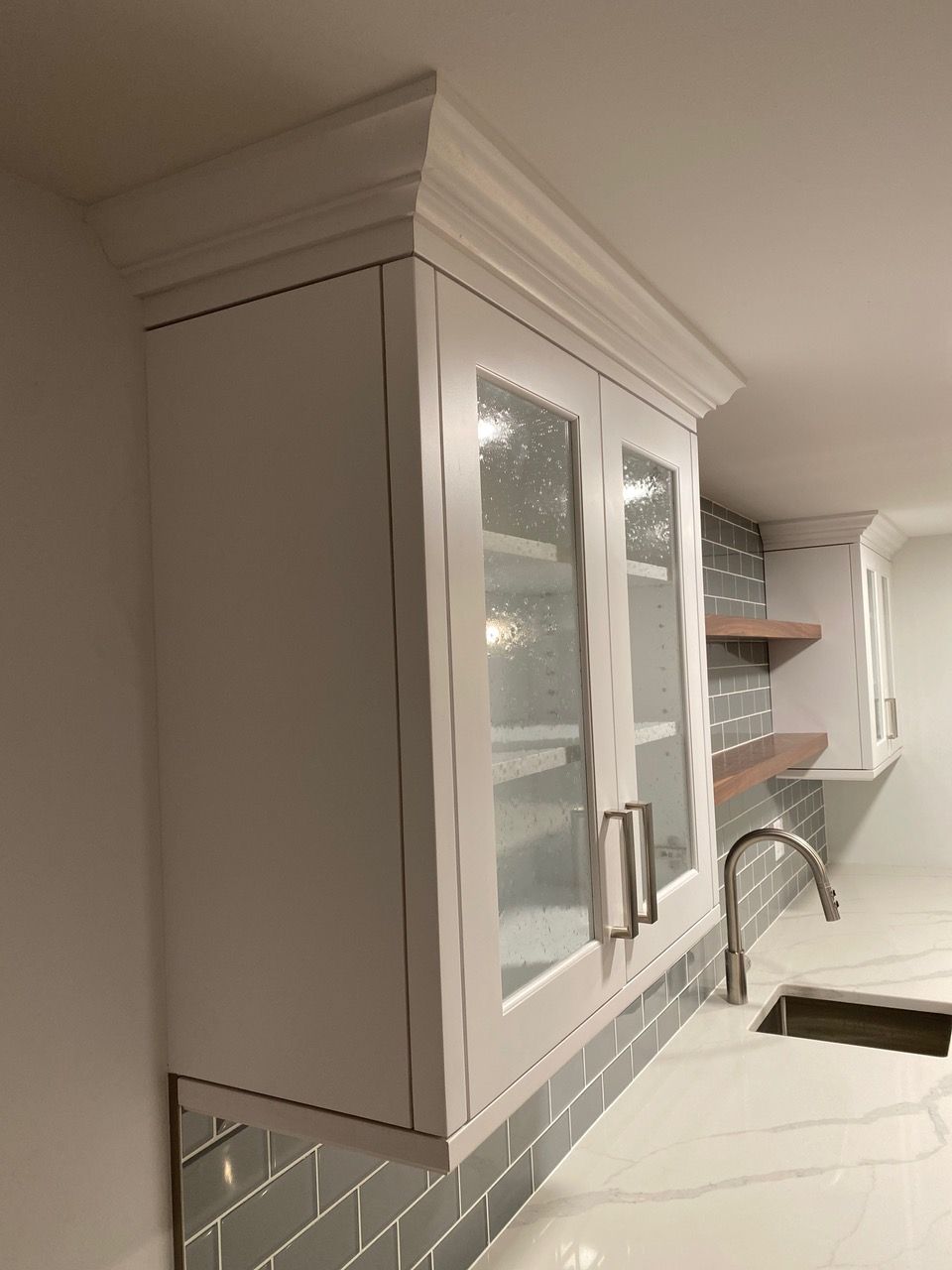White kitchen cabinet with glass doors, mounted above a countertop with a backsplash and sink.