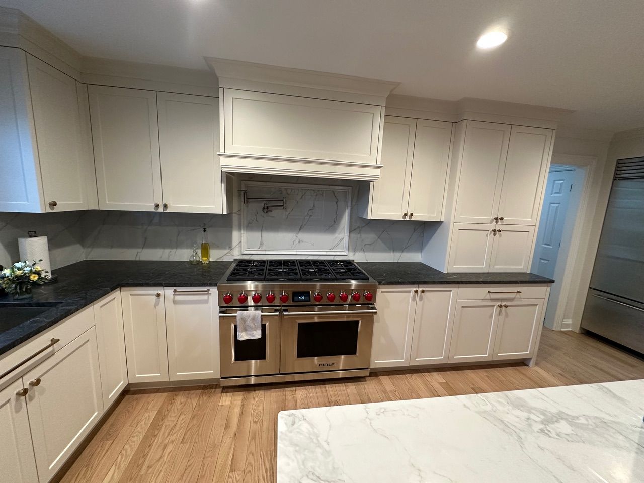 Cream-colored kitchen with stainless steel stove, black countertops, wooden floor, and white cabinetry.