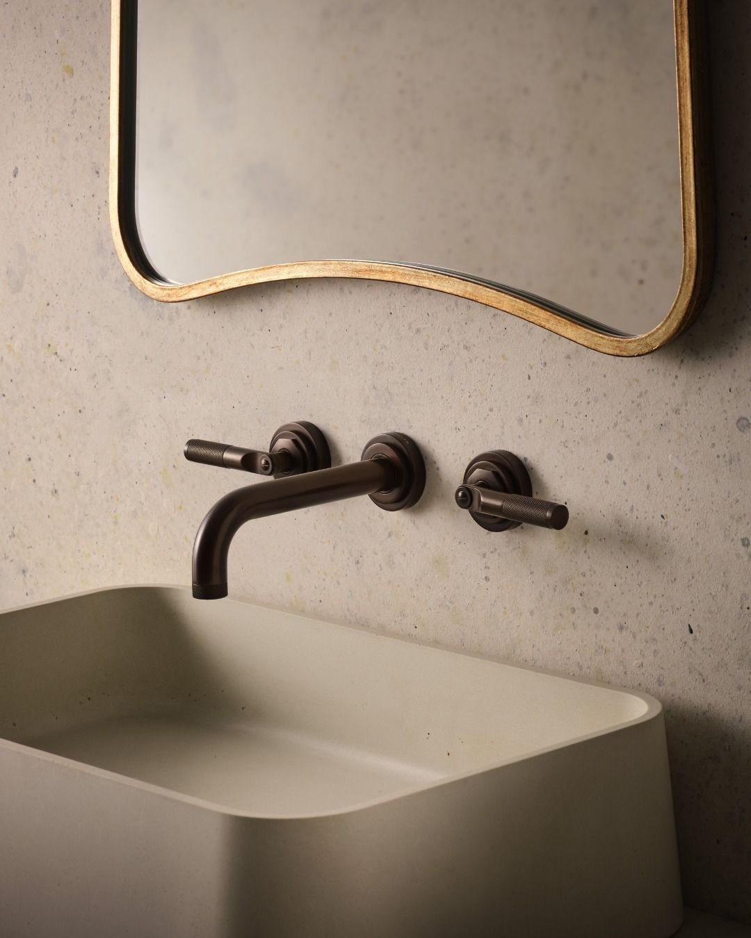 Bathroom sink with dark faucet, handles, and gold-framed mirror against a concrete wall.