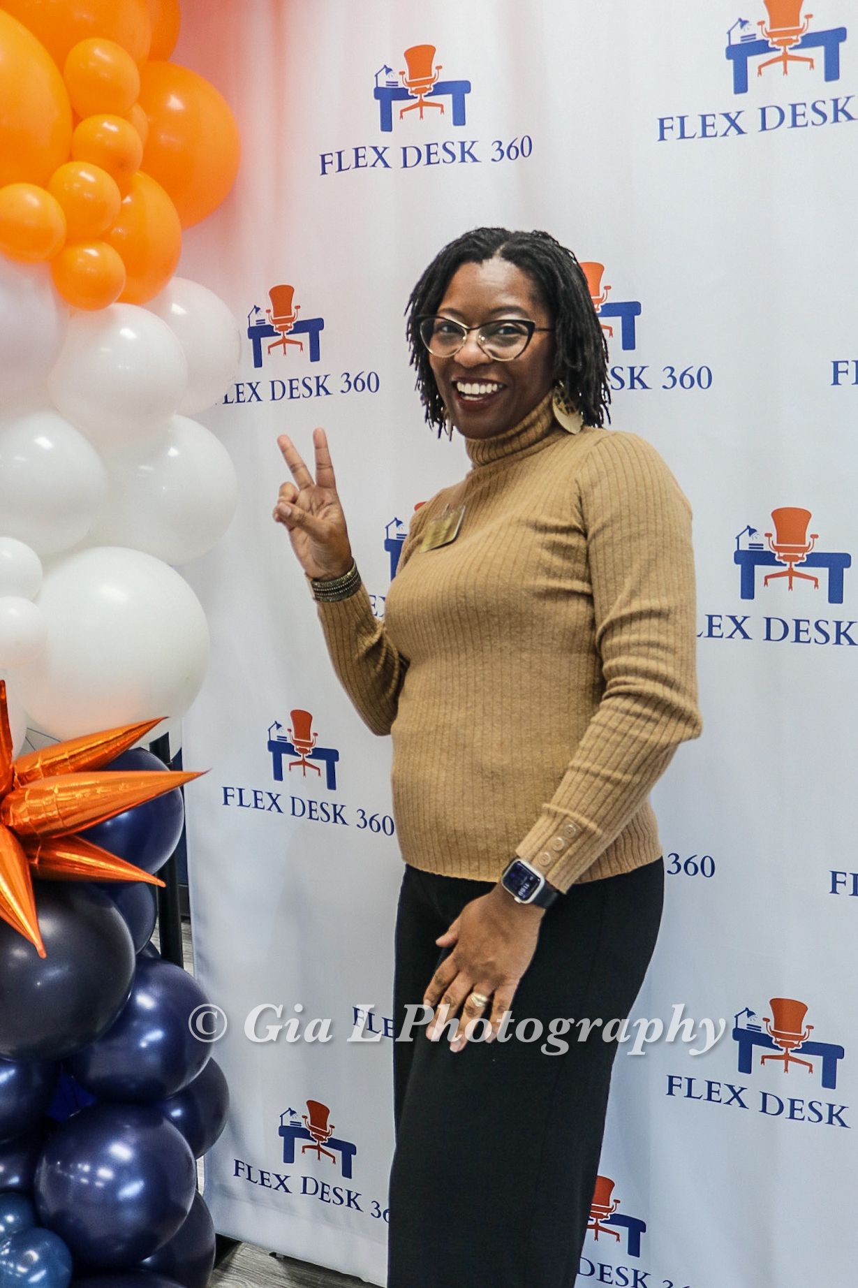 A woman is standing in front of a wall with balloons and giving a peace sign.