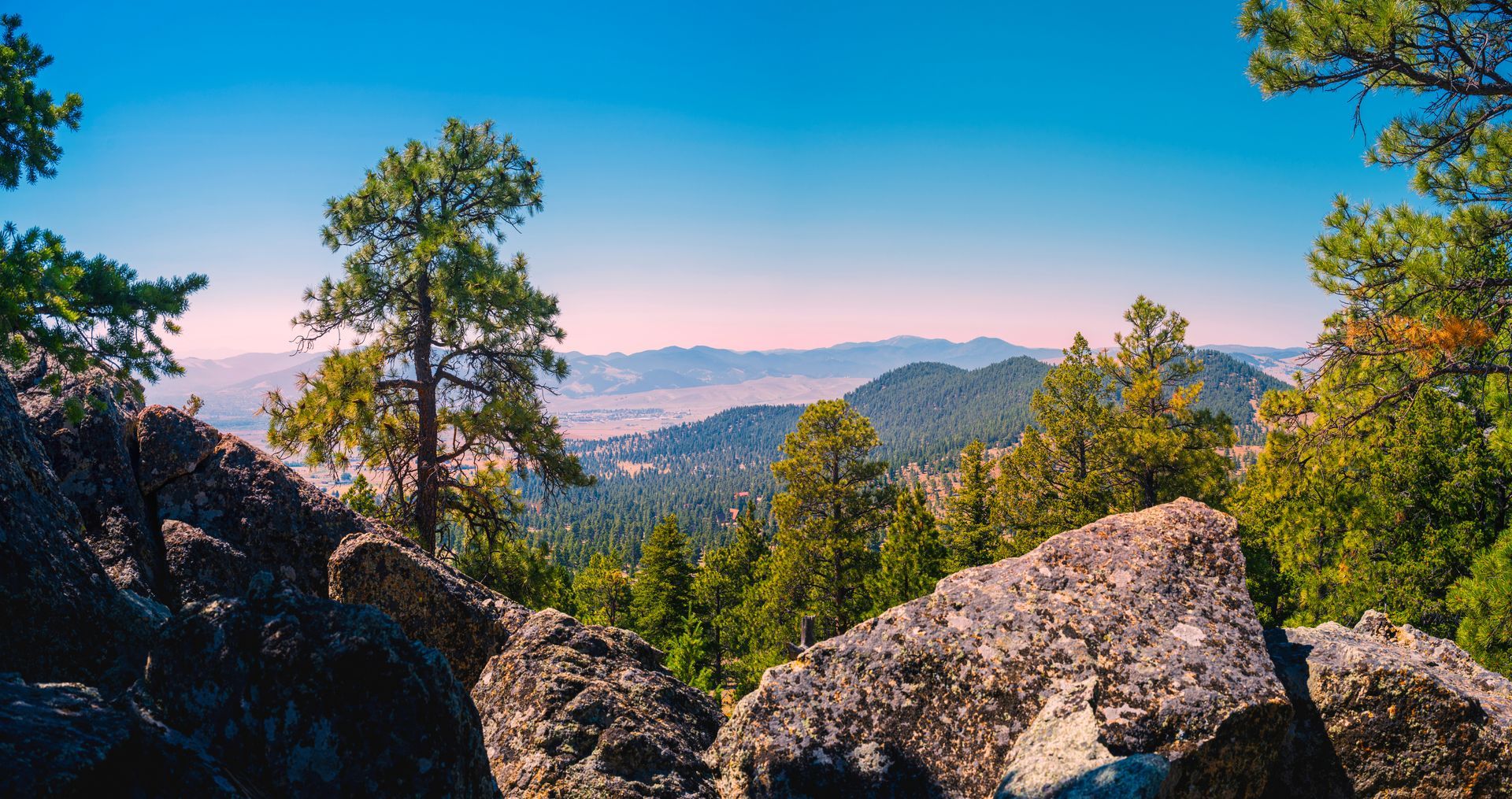 A view of a mountain range from a rocky cliff with trees in the foreground.