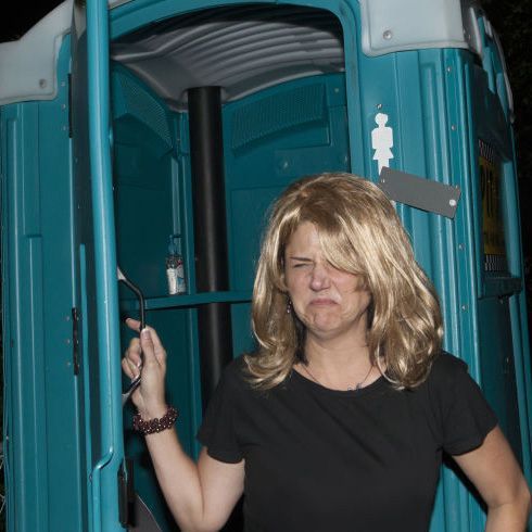 A woman in a black shirt is standing in front of a portable toilet with a disgusted face