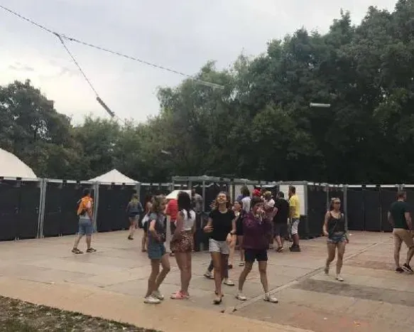 A group of people are standing on a sidewalk in a park. near sanitrax aeroflush portable toilets at a festival