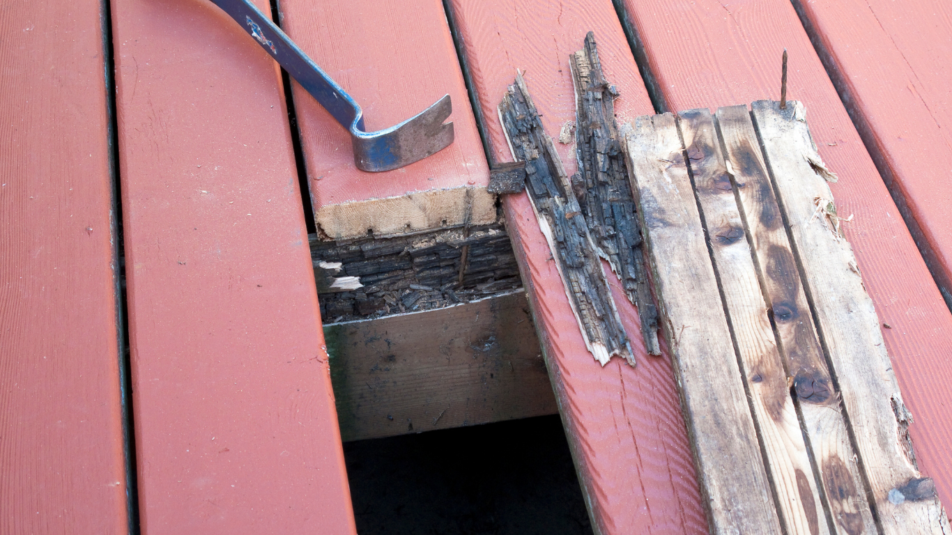 Close-up of deck board being pried up, revealing damage and a dark opening. A crowbar is on the red deck.