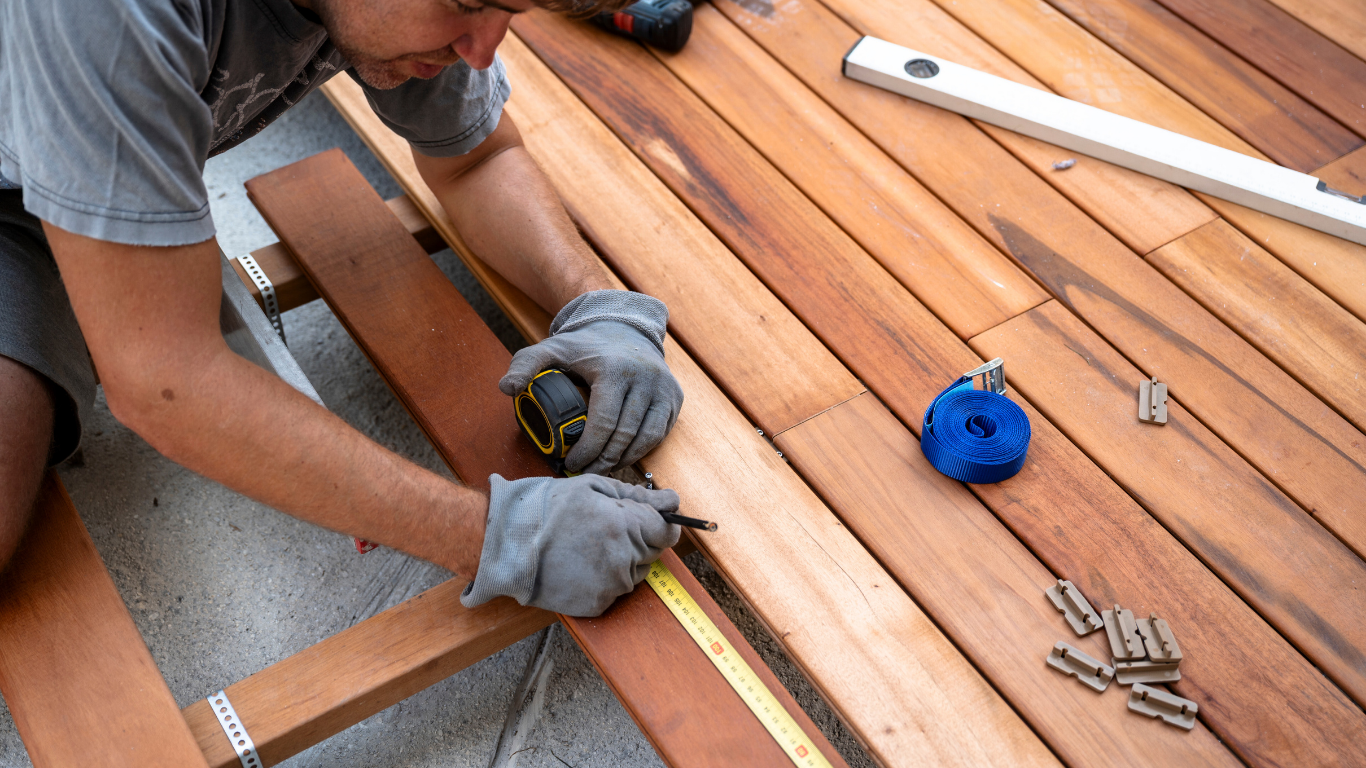 Person measuring and marking a wooden deck with a tape measure, wearing gloves.