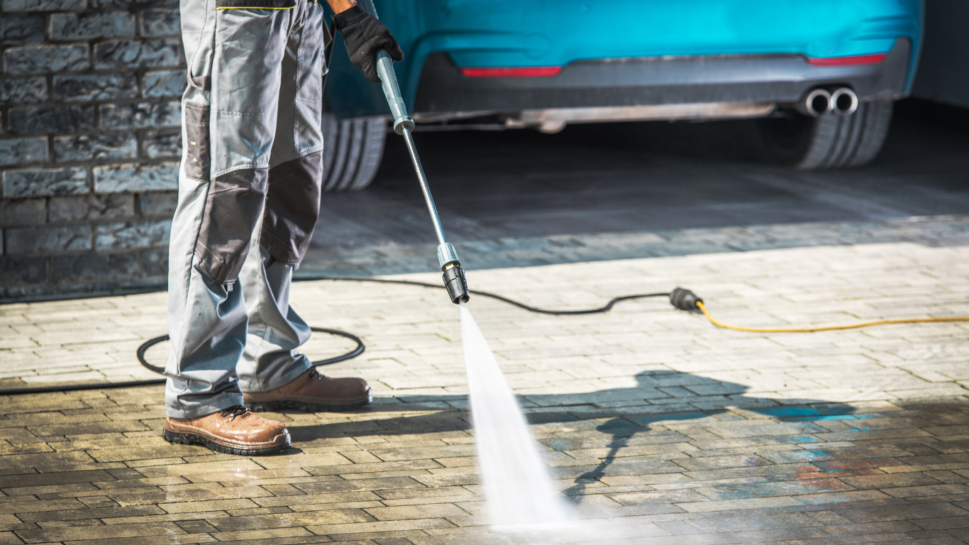 Person power washing a paved driveway next to a teal car.