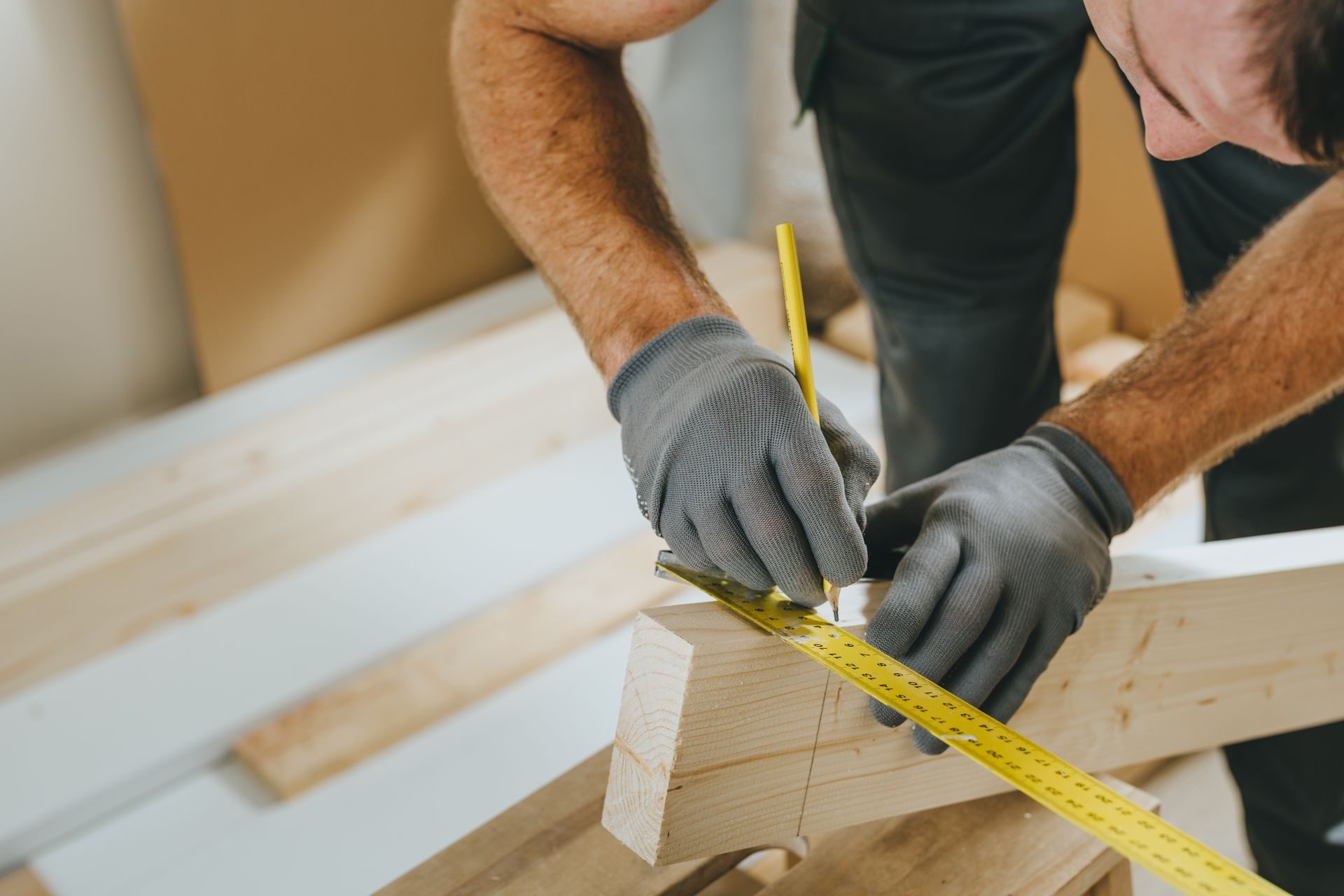 Person in work gloves measuring and marking wood with a pencil and measuring tape.