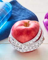 Red apple wrapped with a measuring tape beside blue and purple dumbbells on a white surface
