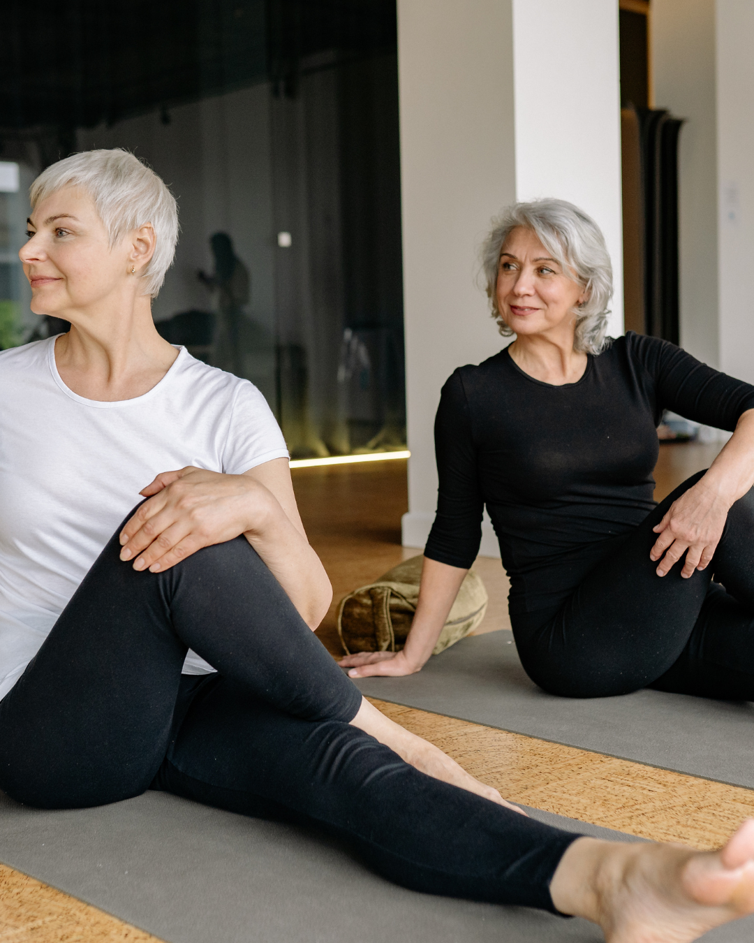 Two people stretching on yoga mats in a bright room, seated and facing different directions.