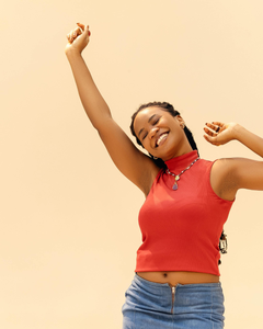 Person dancing with arms raised, smiling in a red sleeveless top and blue jeans against a plain beige background