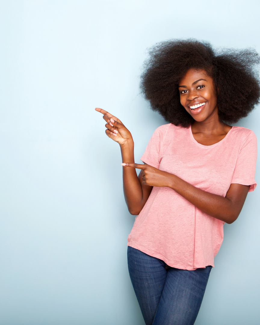 Smiling woman in a pink shirt and jeans pointing to the left against a light blue background