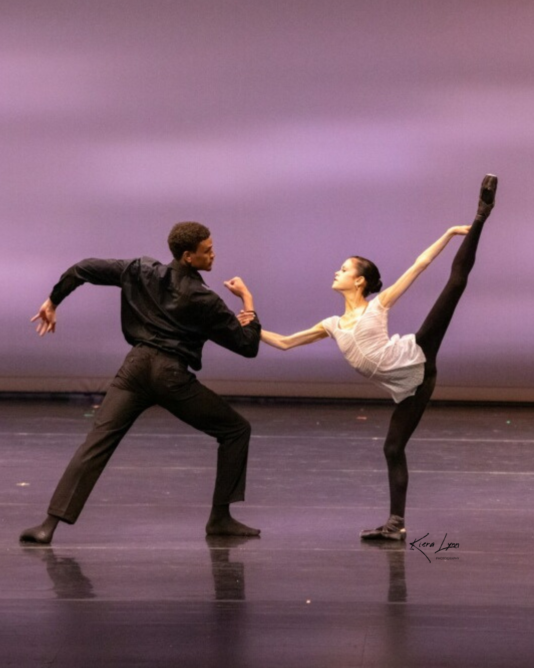 Ballet dancers, man and woman, in black and white outfits on stage, woman in split, purple backdrop.