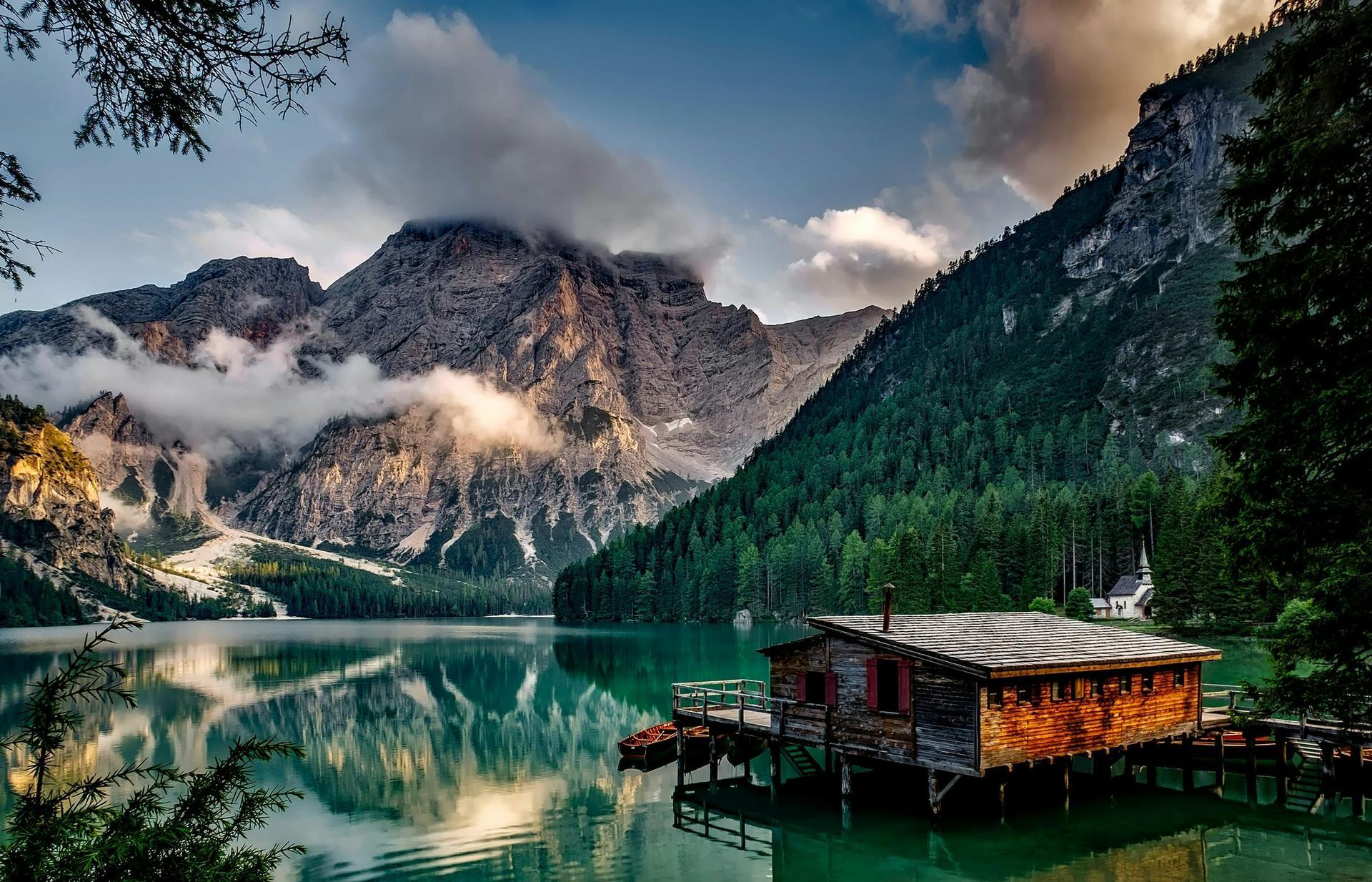 Lakeside cabin, mountains, and trees. Emerald water reflects peaks and clouds under a cloudy sky.