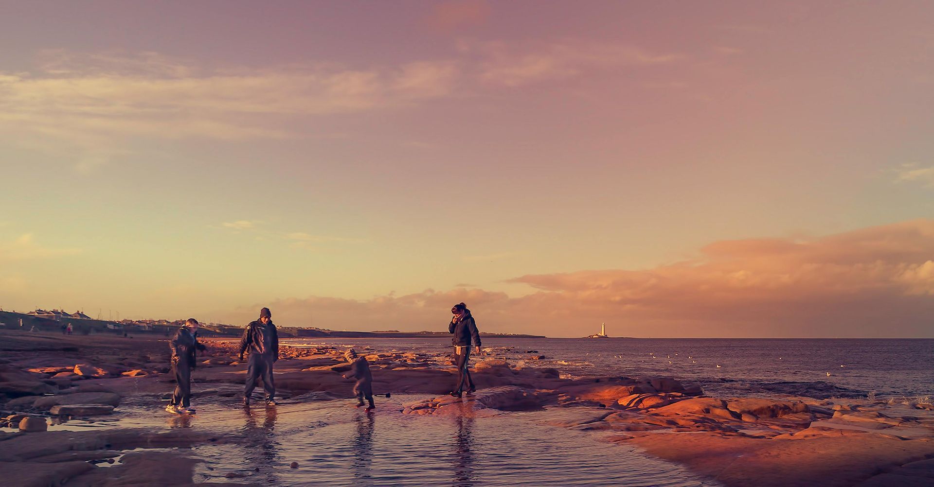 Four people walking on a wet beach at sunset. Sky is orange and pink.