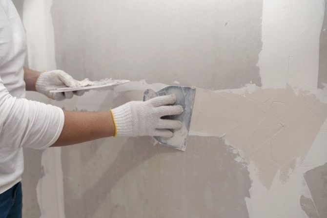 Person smoothing wet plaster on a wall with a trowel while wearing work gloves.