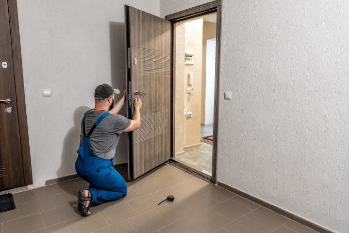 Worker kneeling by a doorway, using a drill to install a wooden door in a tiled room.