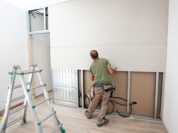 Worker installing drywall in a room with ladders and exposed studs