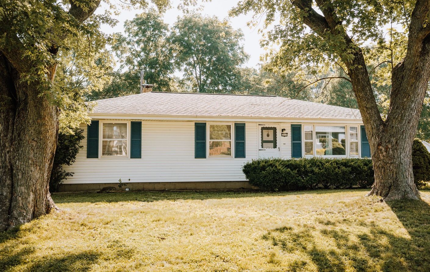 A white house with green shutters is surrounded by trees and grass.