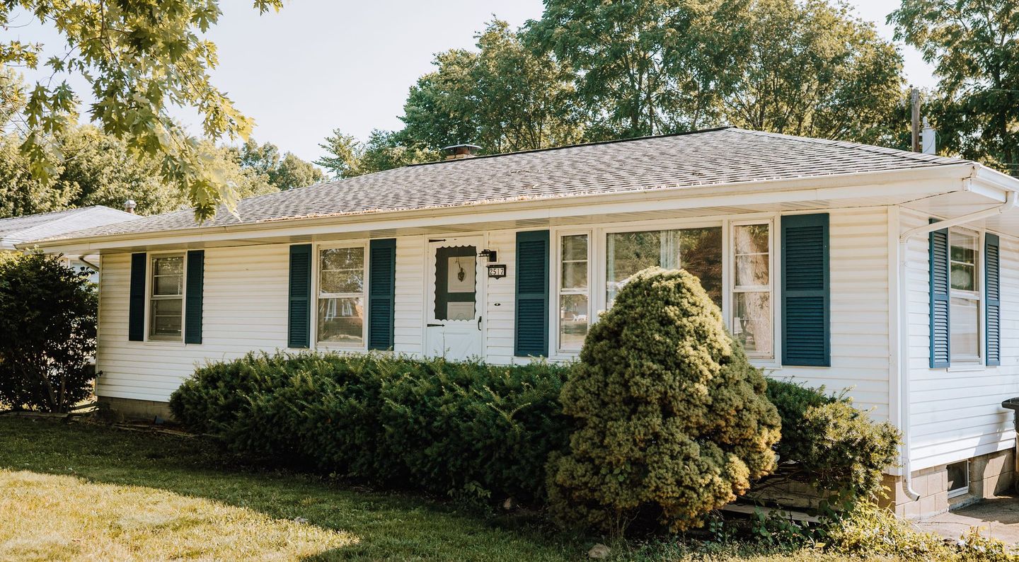 A white house with black shutters is surrounded by trees and bushes.