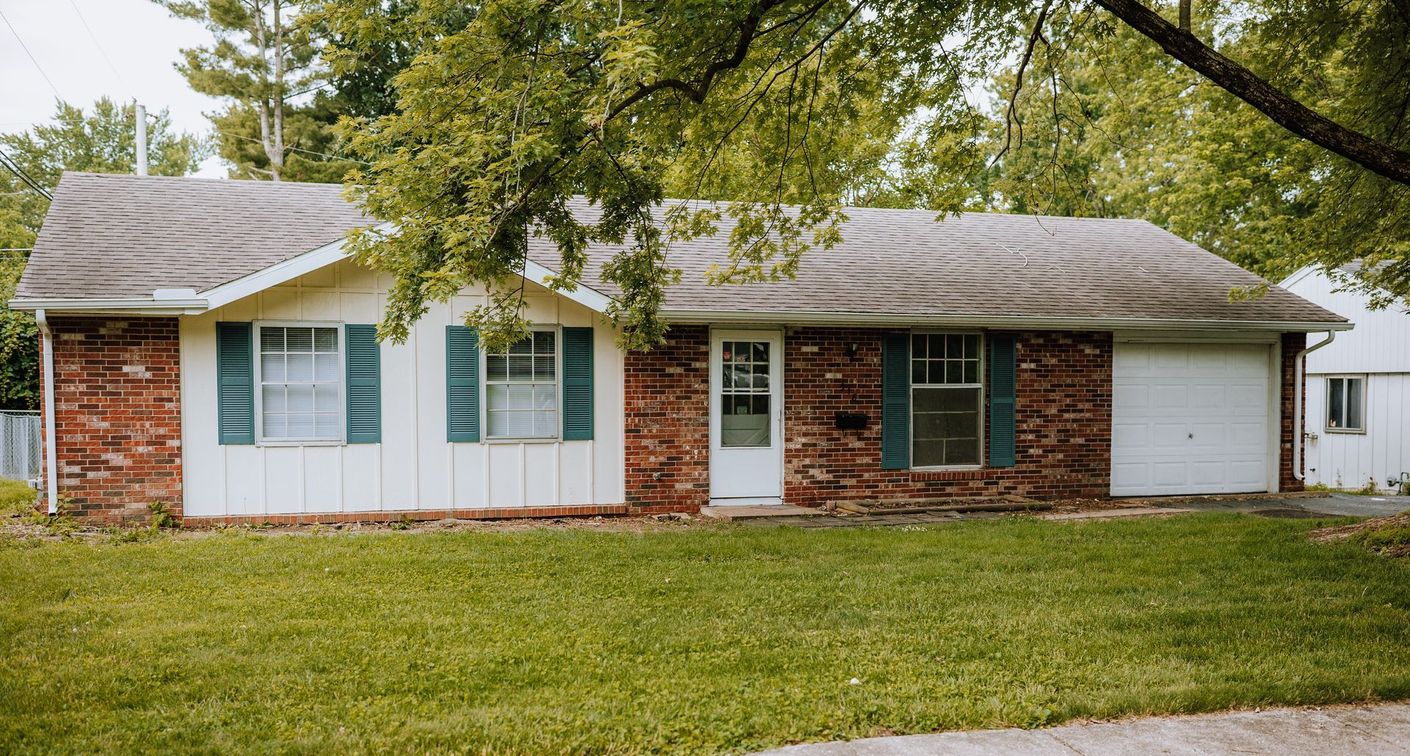 A white house with a brick wall and blue shutters is sitting on top of a lush green lawn.