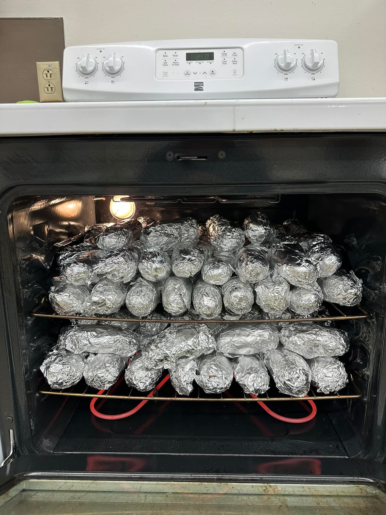 Oven filled with numerous foil-wrapped potatoes on two racks, ready for cooking