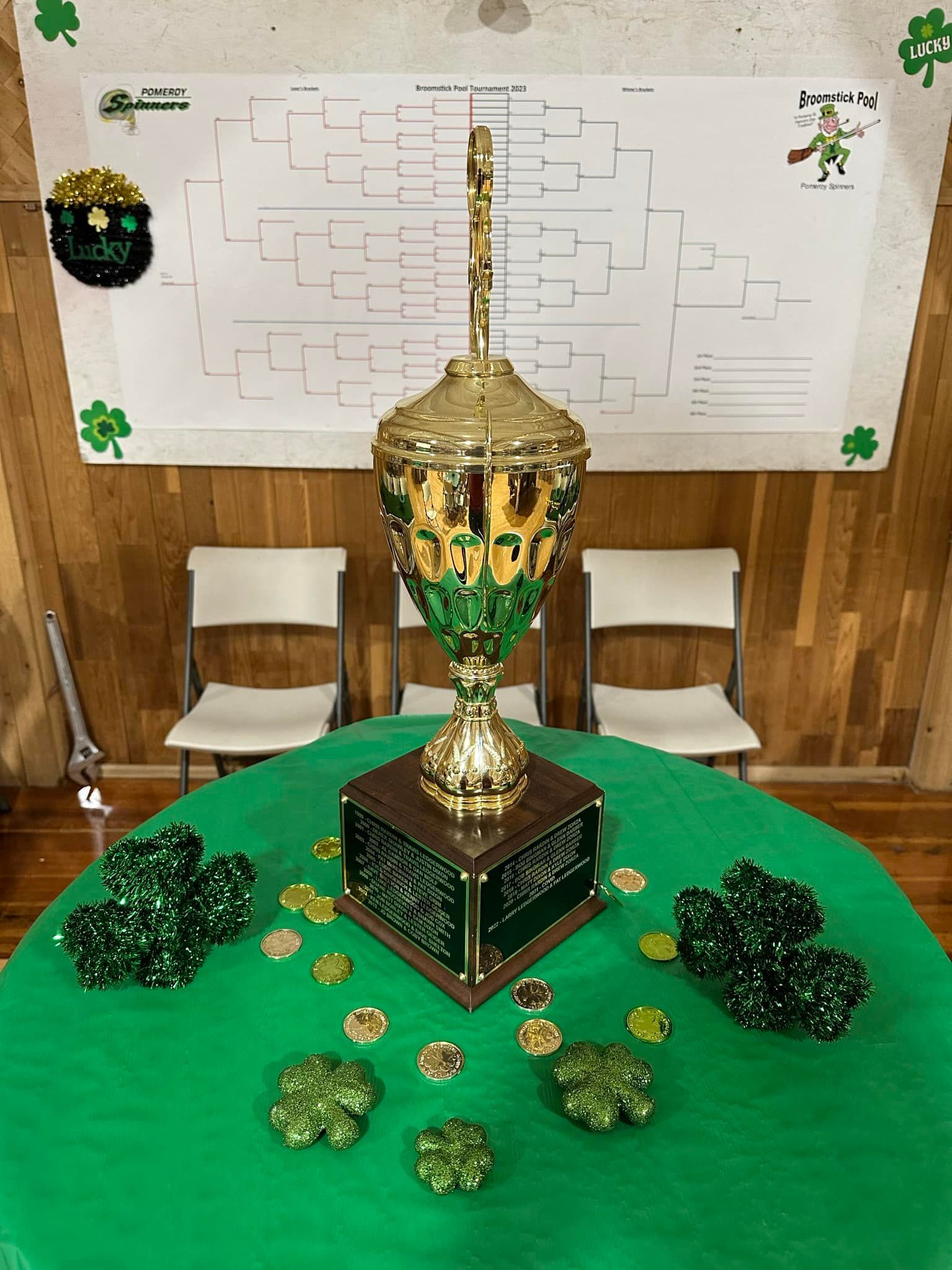 A golden trophy on a green table decorated with shamrocks and coins, with a bracket chart in the background