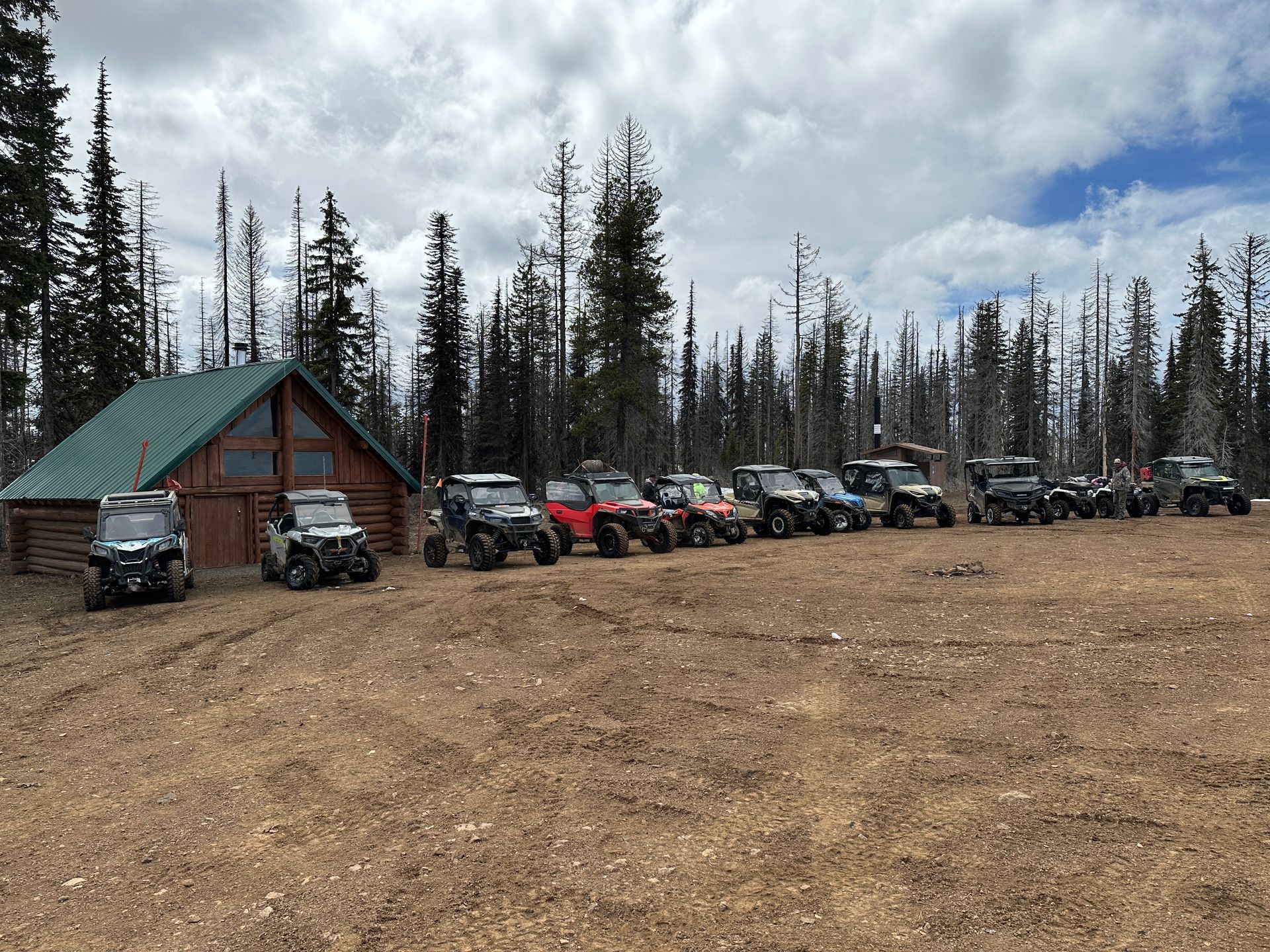 ATVs lined up in a dirt lot in front of a cabin and evergreen trees on a cloudy day.