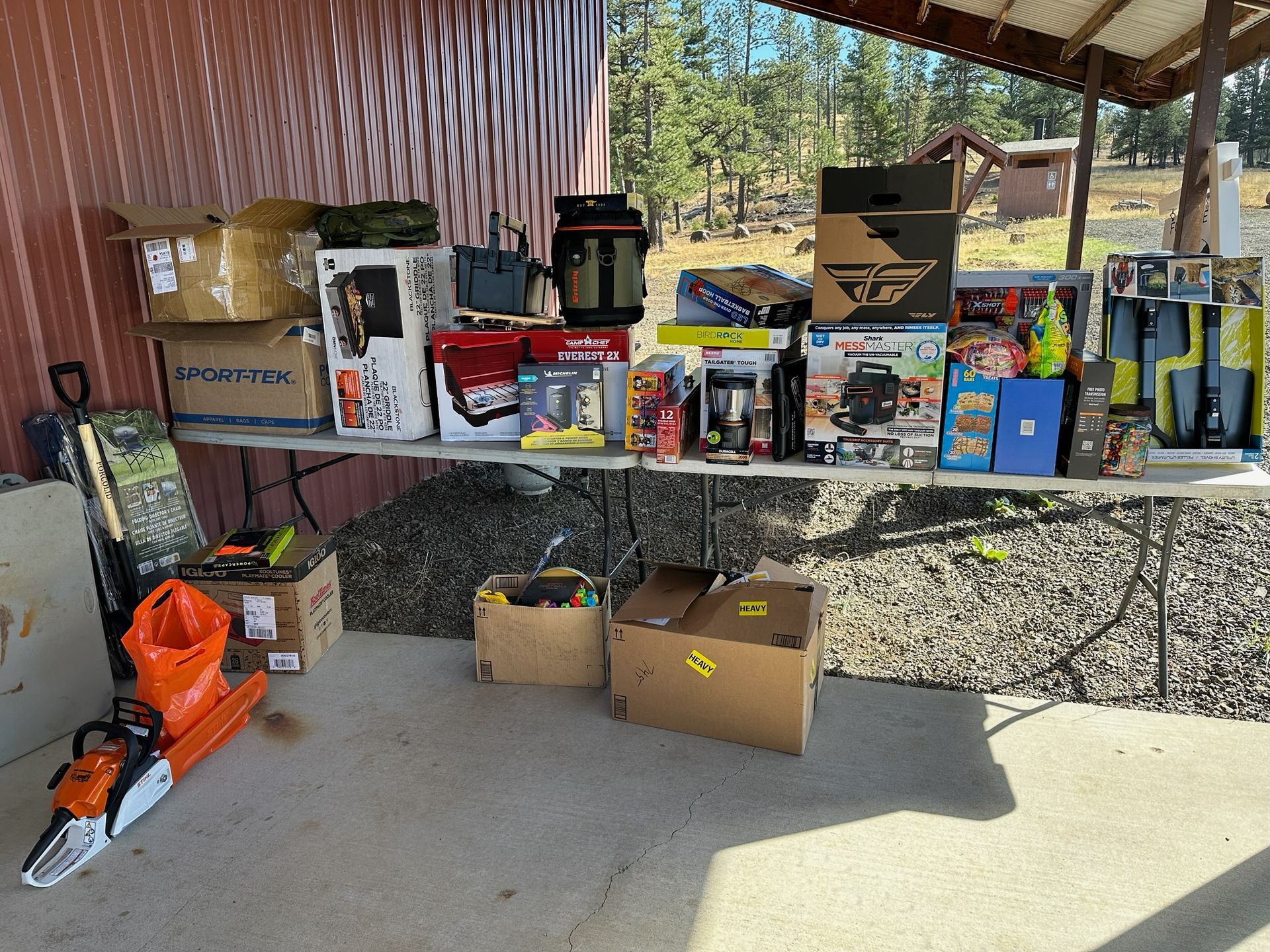 A table displays various items, including tools, electronics, and boxes, outdoors under a shaded structure.