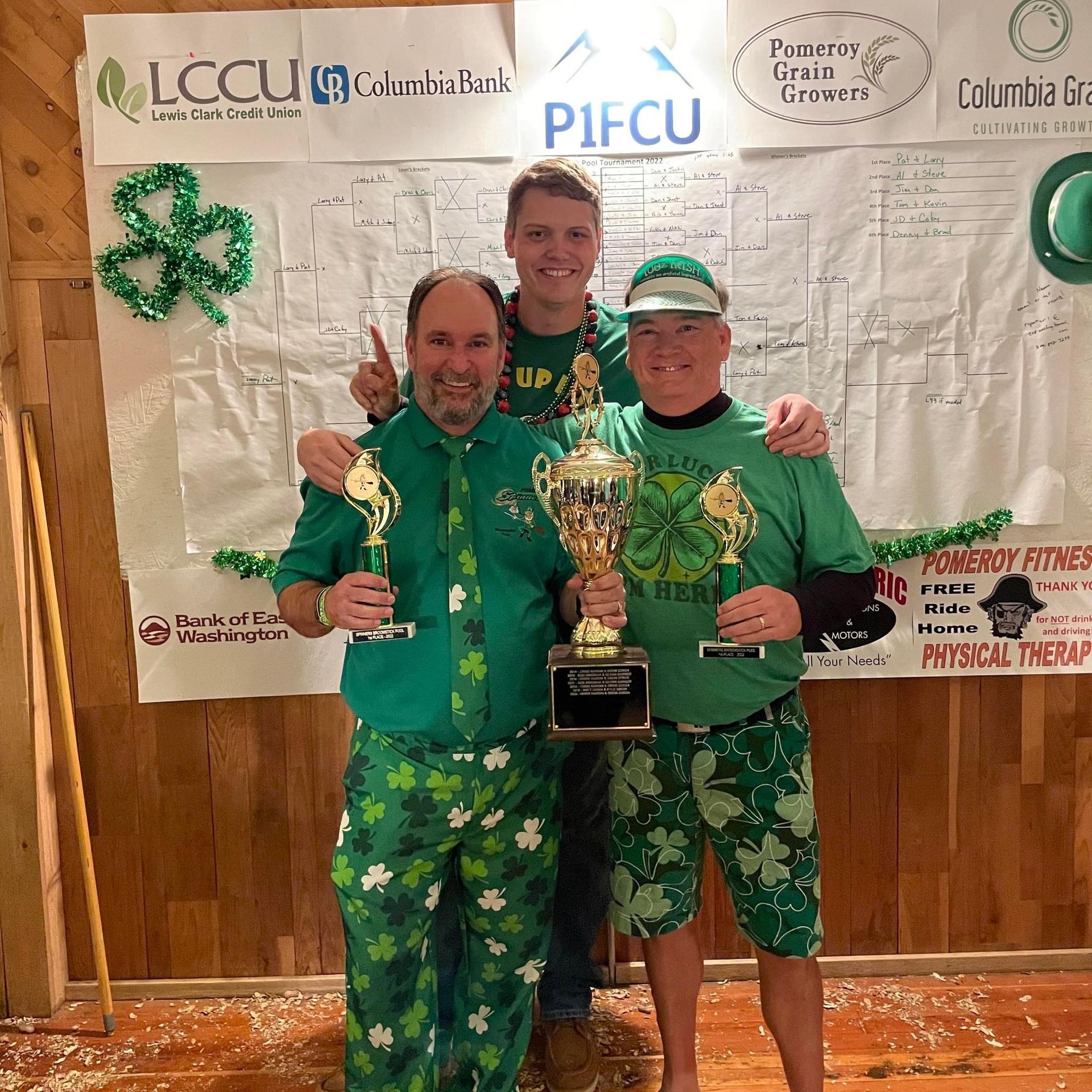 Three men in green St. Patrick's Day attire holding trophies, smiling; festive backdrop with banners.